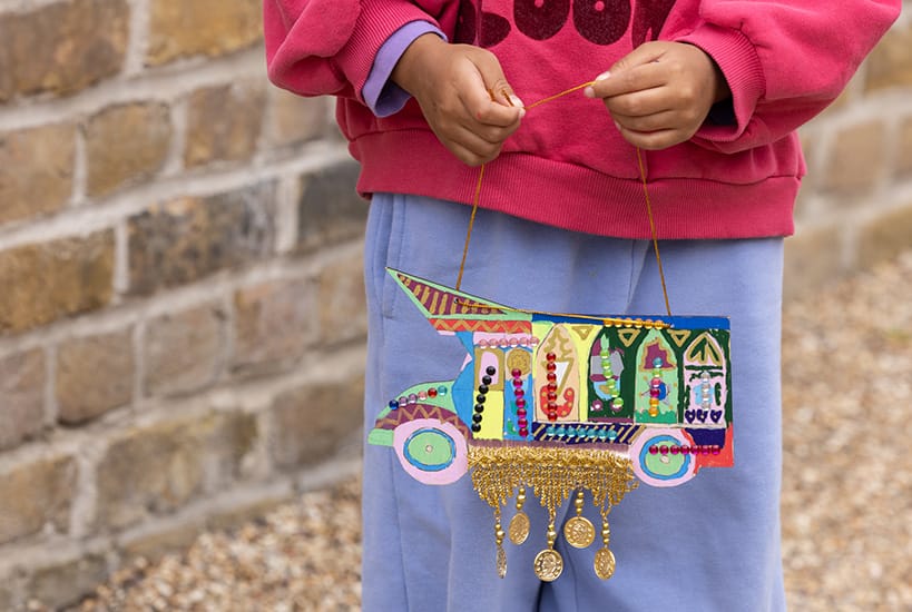 a child holds a small truck-shaped paper craft piece that has been embellished with beads and paint