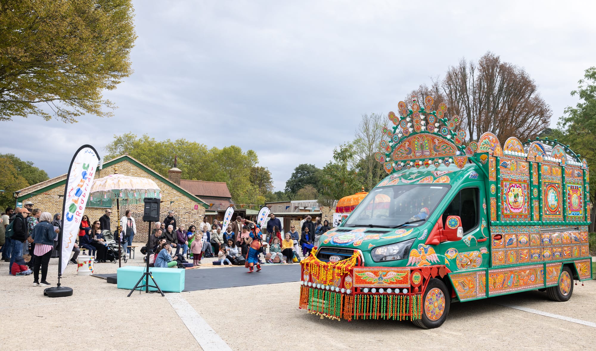 a vibrantly decorated electric van is parked as a background for performances, with a small crowd gathered
