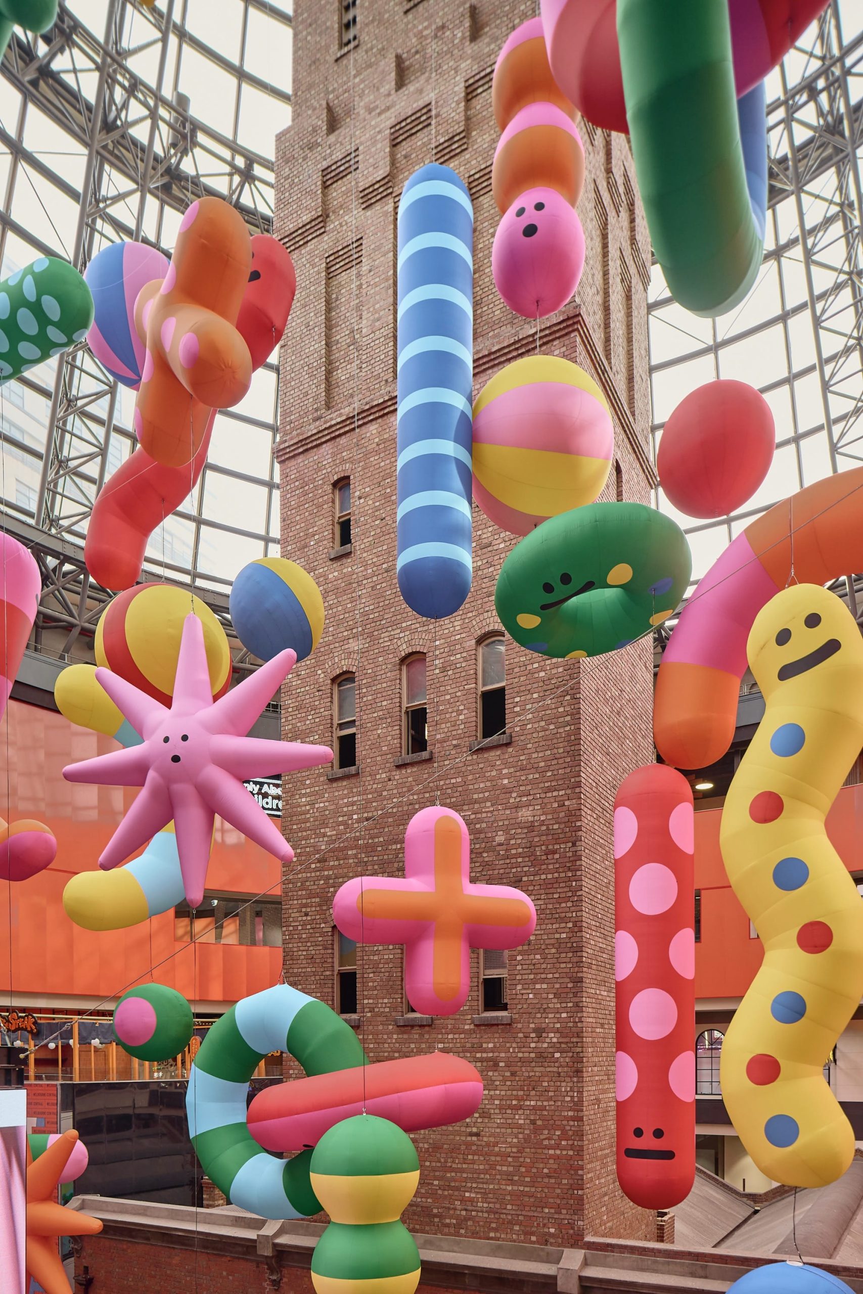 a large-scale installation in Melbourne Central of a number of abstract, colorful shapes, with expressive faces, suspended from the ceiling