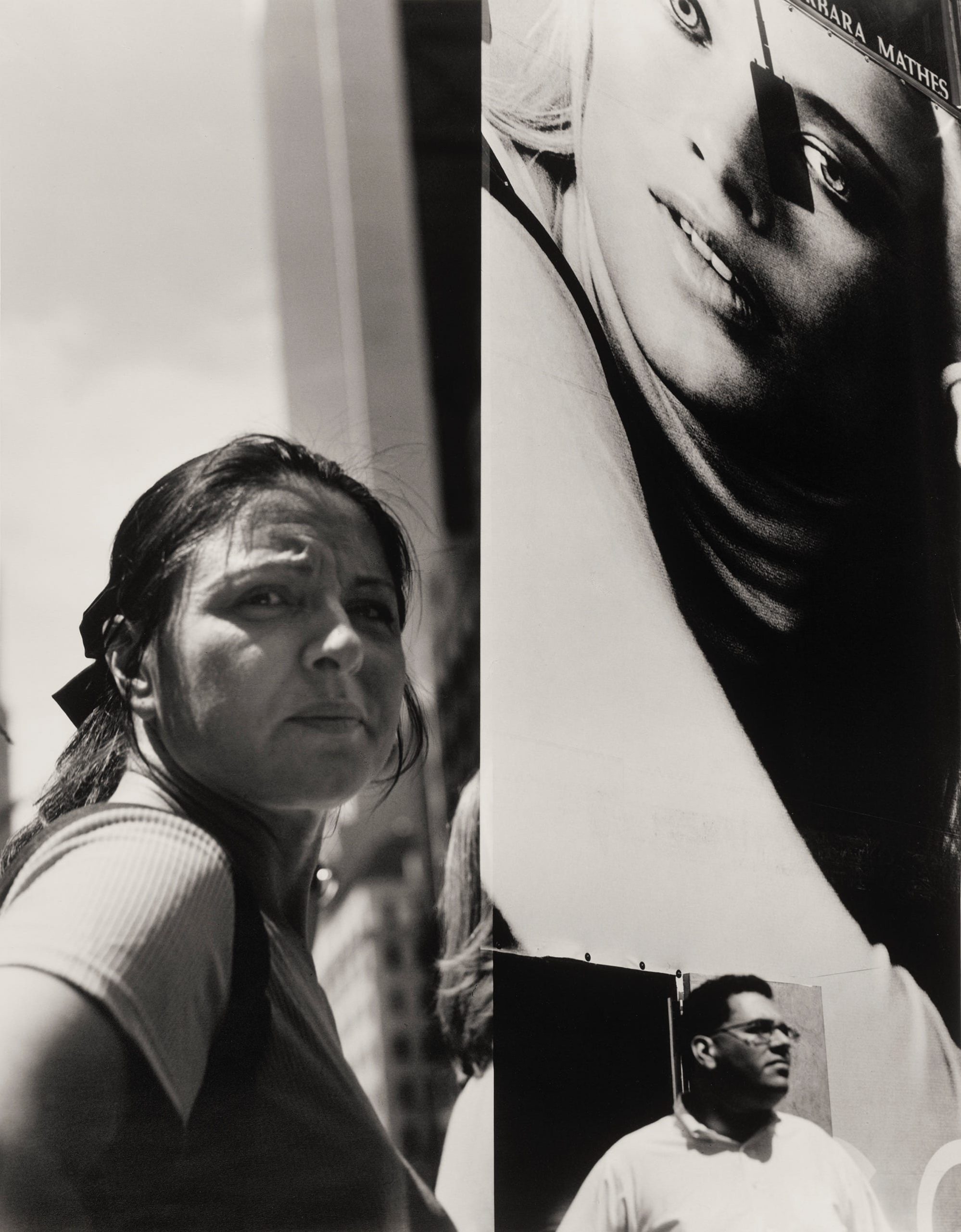 a black-and-white photo by Michael Spano of a woman standing next to an advertisement